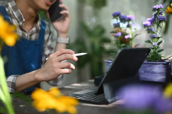 Stock image Pleased male florist talking on mobile phone and using laptop to taking orders at flower shop