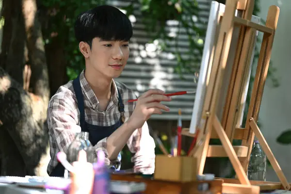 Handsome Asian male wearing apron painting picture with brush on easel in the garden