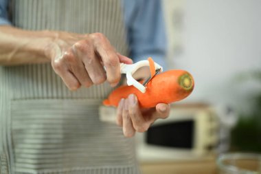 Yakından bakınca önlüklü yaşlı adam mutfakta havuç suyu yapmak için havuç soyuyor.