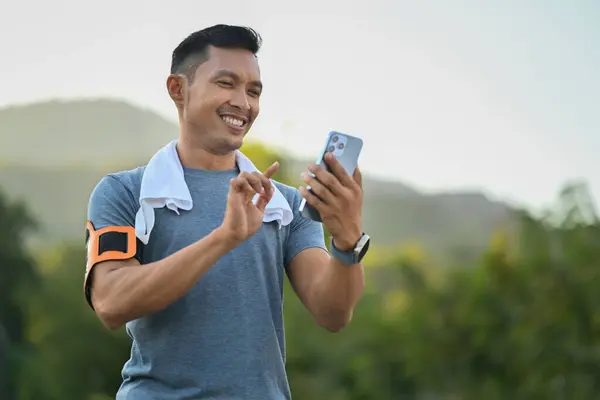 Smiling sporty man checking training results on mobile app after running in the park. Technology health and wellness concept.