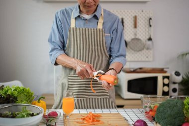 Mutfakta sağlıklı salata yapmak için havuç soyan önlüklü mutlu Asyalı kıdemli adam.