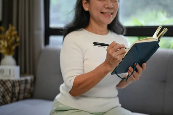 Smiling woman 50s years old wearing casual clothes writing her diary while relaxing at home