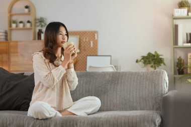 Evdeki kanepede dinlenen sıcak bir fincan kahve tutan mutlu genç kadın.