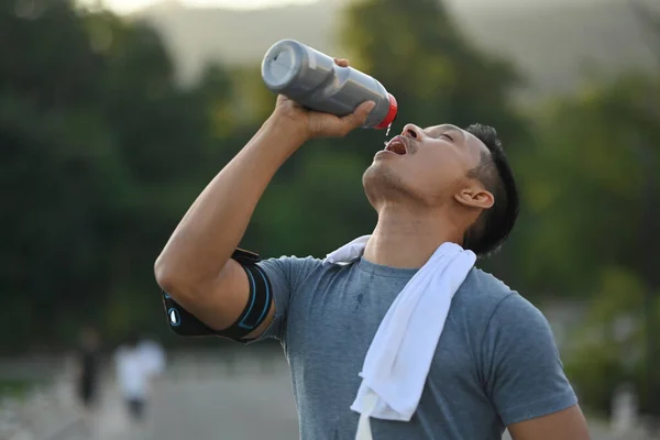 Handsome athletic male drinking water from bottle after running resting after workout outdoors