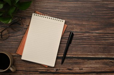 Boş not defteri, bir fincan kahve, kalem bardakları ve ahşap masada saksı çiçeği. Düz yatış, üst görünüm.