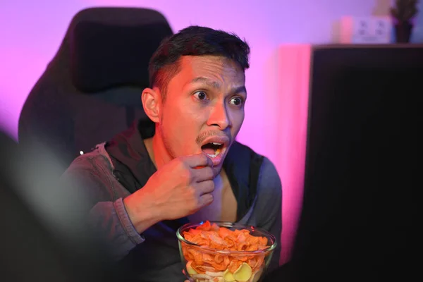 Shocked Asian man with mouth open watching horror movie at home.