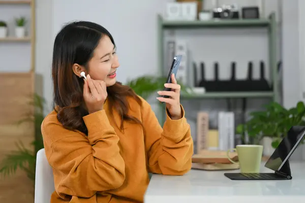 Cheerful young Asian woman freelancer wearing wireless earphones and using mobile phone