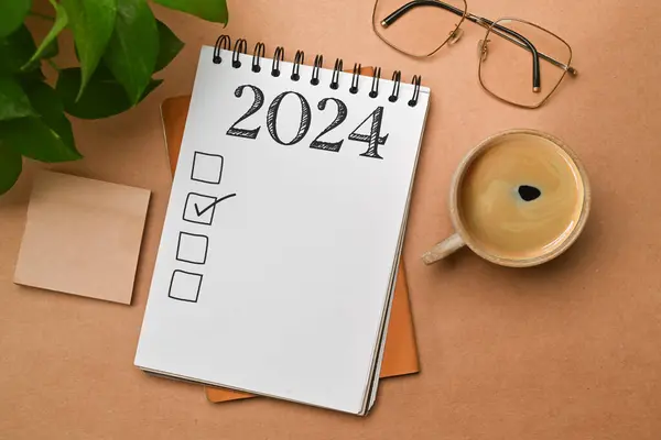 2024 goals list with notebook, coffee cup, plant and glasses on brown background. Plan, journal, to-do list.