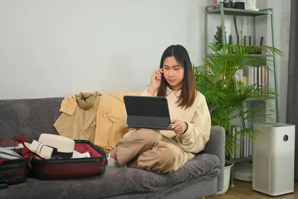 Young woman sitting near unpacked suitcases and using digital tablet for booking hotel online.