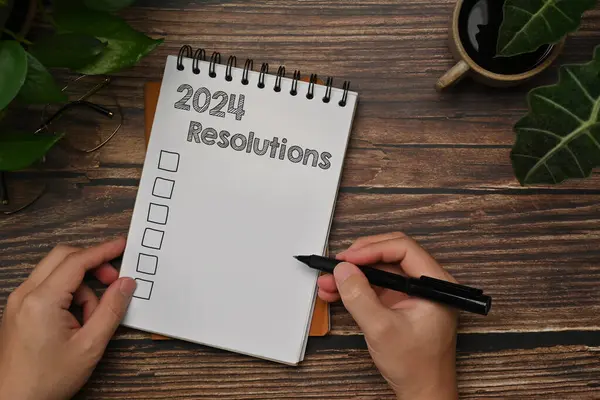 Man hand writing in notebook with list of 2024 resolutions on wooden table. Top view