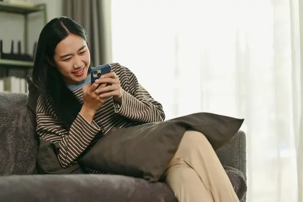 Happy young woman chatting with friend on mobile phone while relaxing on couch.