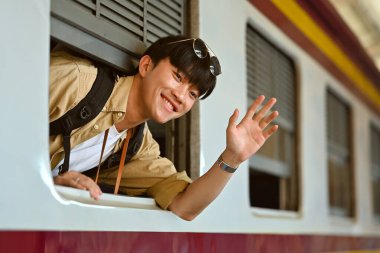 Tren penceresinden el sallayan mutlu Asyalı erkek sırt çantalı. Ulaşım, insanlar ve seyahat kavramı