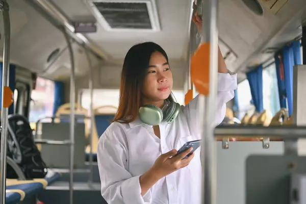 Attractive young woman standing in bus and using smart phone during her morning commute.