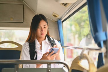 Güneşli bir günde otobüste seyahat ederken cep telefonu kullanan sırt çantalı genç bir kadın..