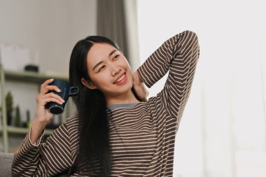 Rahat genç bir kadın, elinde kahve fincanı, uzayan bir vücut, rahat bir evde, kanepede dinleniyor..