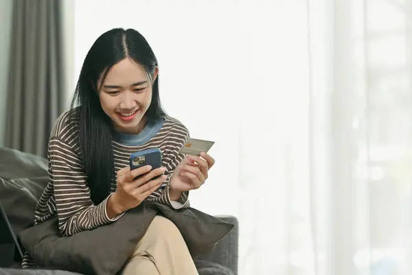 Charming young Asian woman holding credit card and doing online banking transaction on mobile phone.
