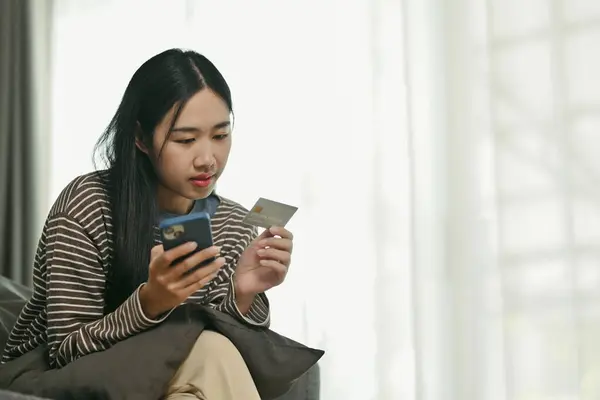 Young Asian woman holding credit card and doing online banking transaction on mobile phone.
