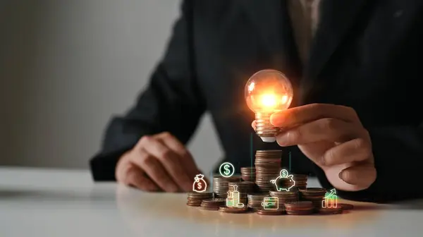Businesswoman hand holding glowing light bulb on coin stacking. Saving money and financial growth concept.