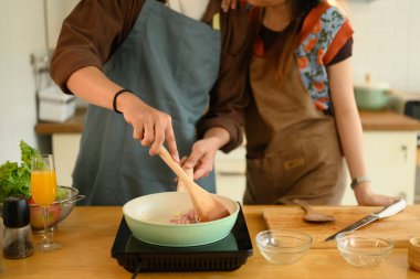 Önlüklü genç çift birlikte yemek hazırlıyor, spatula kullanan adam kıymalı domuz ve sarımsağı tavada karıştırıyor..