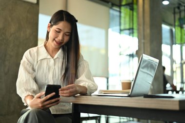 Uzaktan kumandalı kahve dükkanında bilgisayarla otururken cep telefonu kullanan gülümseyen bir iş kadını..