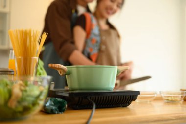 Mutfaktaki elektrikli ocağın üzerinde metal bir tencere. Arka planda genç bir çift yemek pişiriyor..