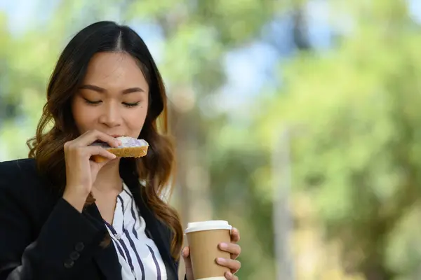 Millennial woman in business suit drinking coffee and eating sandwich in the park.