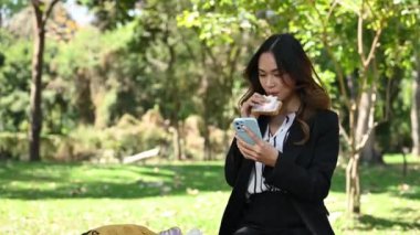 Güzel genç iş kadını sandviç yiyor ve parkta cep telefonu kullanıyor..