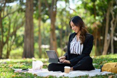 Parktaki çimlerin üzerinde oturan dijital tablet üzerinde çalışan büyüleyici bir iş kadını..