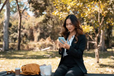 Tahta bir bankta oturan ve cep telefonu kullanan çekici kadın ofis çalışanı..