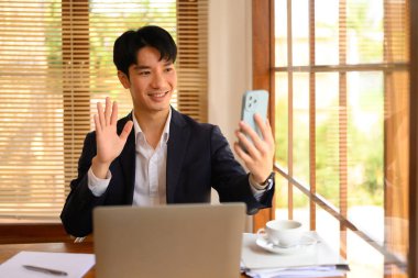 Gülümseyen işadamı çalışma masasında oturuyor ve cep telefonuyla görüntülü konuşuyor..