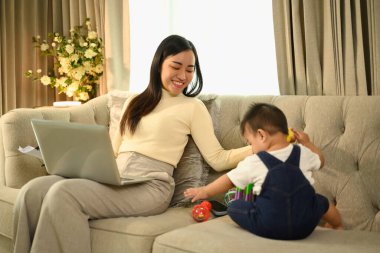 Genç, güzel, sevgi dolu anne bebeğiyle ilgilenirken dizüstü bilgisayarda çalışıyor..