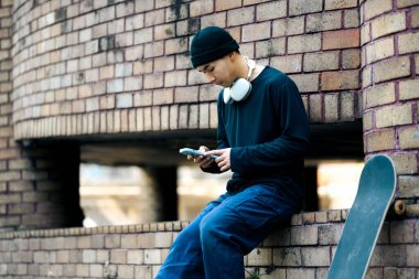 Şehir caddesinde kaykayla otururken cep telefonu kullanan şık giyimli yakışıklı Asyalı adam..