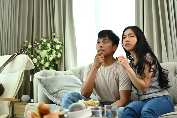 Excited young couple watching scary horror movie during weekend in living room.