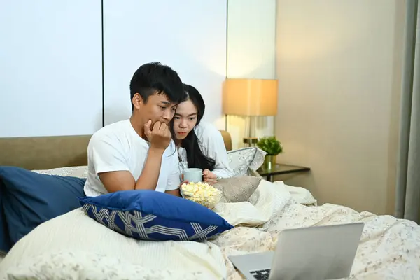 Excited couple watching horror movie on digital tablet during weekend in bedroom.