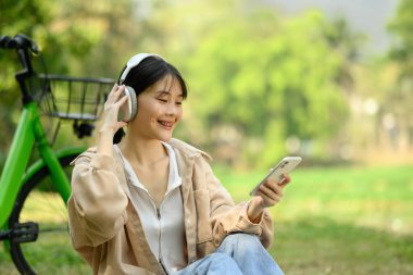Parkta bisikletle otururken cep telefonu ve kulaklıkla müzik dinleyen güzel bir genç kadın..