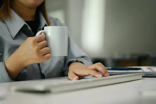 Cropped shot of female office worker holding coffee mug and typing on computer keyboard.