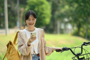 Sırt çantalı gülümseyen genç bir kadın bisiklet parkında duruyor ve cep telefonuyla sohbet ediyor..