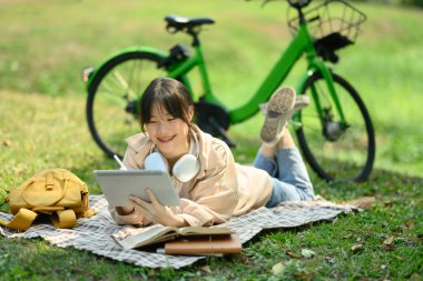 Kamu parkında battaniyeye uzanmış gülümseyen genç bir kadın ve dijital tablet kullanıyor..