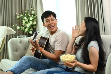 Kız arkadaşı için gitar çalan genç bir adamın evdeki kanepede oturması..