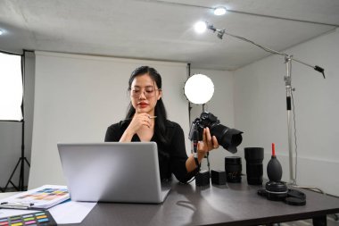 DSLR kamerasını tutan yaratıcı genç bir kadın ve fotoğraf stüdyosunun iç kısmında dizüstü bilgisayarla çalışıyor..