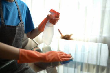 Kadın eldiven giyip cam masayı bez ve dezenfektanla temizliyor..