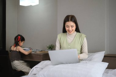 Güzel anne dizüstü bilgisayarda çalışıyor ve anaokulu kızı yatak odasında ödev yapıyor..