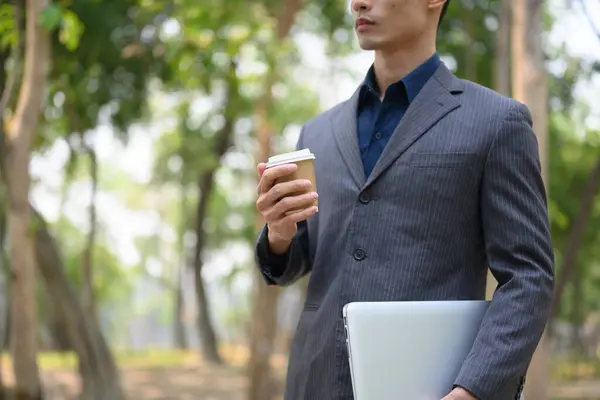 Millennial businessman drinking takeaway coffee during walking in the public park.