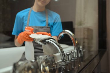 Temizlikçi, banyo musluğunu bez ve deterjan spreyiyle temizliyor. Temizlik hizmeti kavramı.