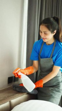 Asyalı kadın temizlikçi sprey ve bez ile yatak başlığını dikkatlice temizliyor..