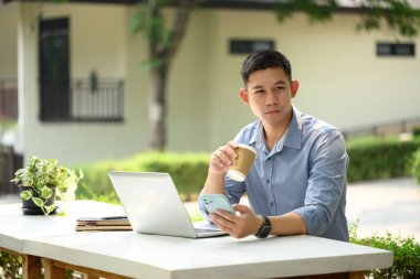 Cep telefonu kullanan ve açık havada kahve içen Asyalı erkek girişimcinin portresi.