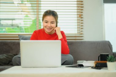 Kanepede oturan neşeli genç Asyalı kadın dizüstü bilgisayarla evde çalışıyor..