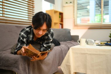 Mutlu genç adam kanepede uzanıp kitap okuyor. Eğlence ve eğlence etkinliği kavramları.