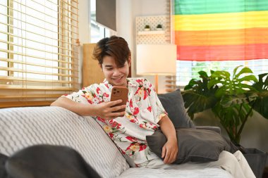 Oturma odasında gökkuşağı LGBT gurur bayrağıyla otururken cep telefonu kullanan gülümseyen genç adam..