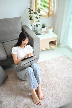 Oturma odasında dijital tablet kullanan mutlu genç bir kadın..